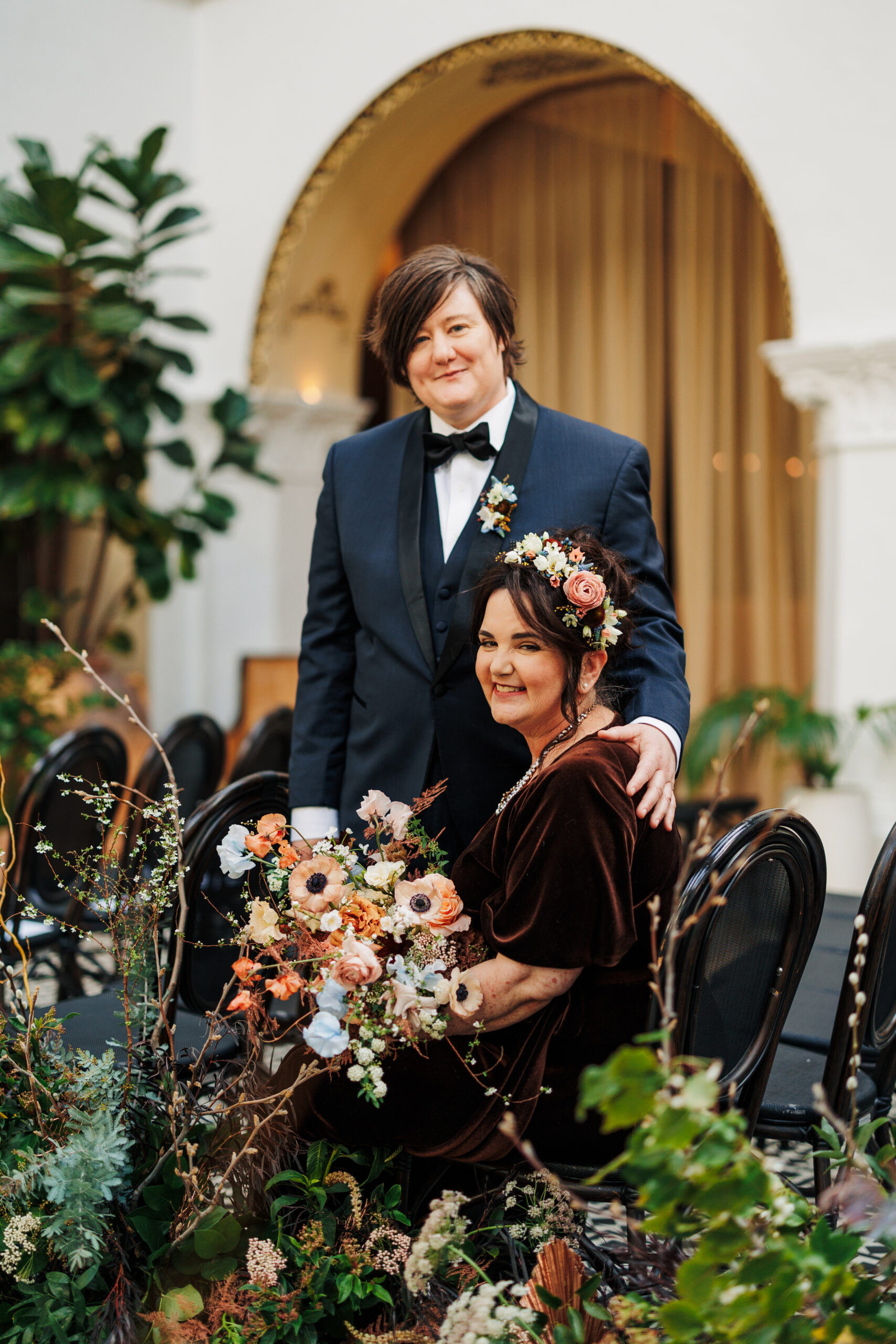 Two women, one sitting one standing surrounded by flowers on their wedding day.